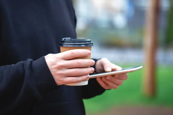 stock image A man holds a paper cup and a smartphone in his hands on a blurred background, Gadget close-up, Disposable paper cup.