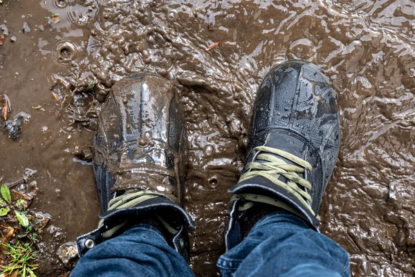 View from above on pair of trekking shoes in a mud, a man in jeans and boots walks through the swamp in rainy weather, close up.