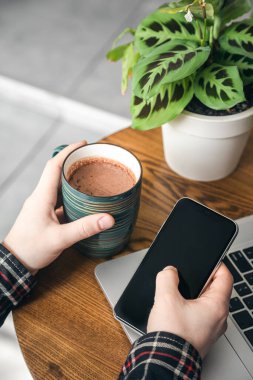 Bilgisayarın önündeki adamın elinde bir fincan kakao ve bir akıllı telefon. Ofiste çalışan bir adam, yakın plan..