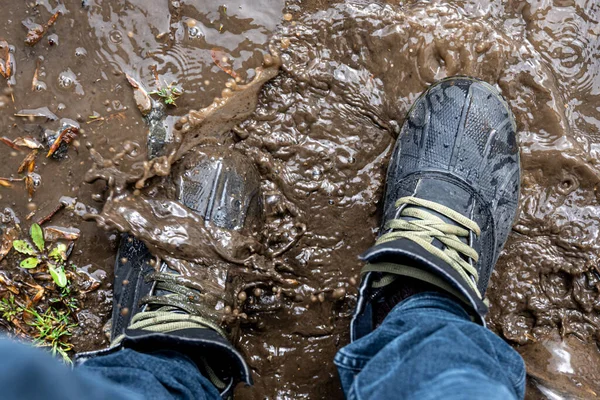 View from above on pair of trekking shoes in a mud, a man in jeans and boots walks through the swamp in rainy weather, close up.