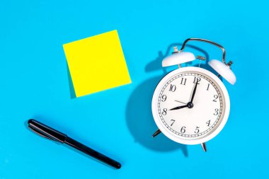 Mavi zemin üzerinde beyaz alarm saati ve boş sarı kağıt etiket izole, düz döşeme, kopyalama alanı.