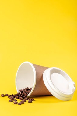 Tek kullanımlık kahve fincanı ve dağınık kahve çekirdekleri sarı arka planda izole edilmiş, kopyalama alanı.