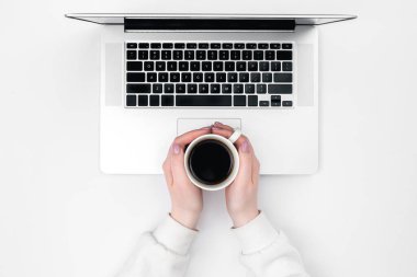 Kadın ellerinde bir fincan sade kahve ve beyaz arka planda bir laptop, üst manzara. Minimalist işyeri.