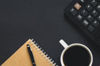 Hesap makinesi, kahve fincanı ve siyah arkaplan üzerine not defteri, minimalist çalışma ortamı, kopyalama alanı.