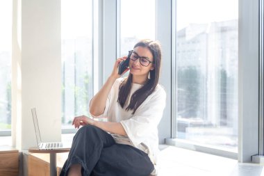 Gözlüklü neşeli genç bir kadın ofisin arka planında akıllı bir telefonla konuşuyor. Çalışma konsepti..