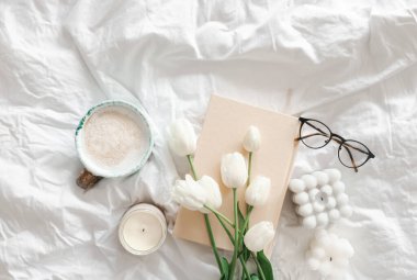 Bir buket beyaz lale, bir fincan kahve, mumlar ve beyaz bir yatakta bir kitap..