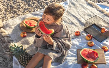 A little girl on a sandy seashore on a blanket at a picnic with fruits eats watermelon.