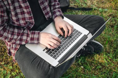 Parkta elinde dizüstü bilgisayarla çimlerin üzerinde oturan bir adam. Kapalı elleri ve laptopu var. Yazın dışarıda çalışıyor..