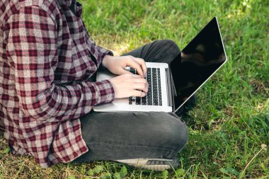 Parkta elinde dizüstü bilgisayarla çimlerin üzerinde oturan bir adam. Kapalı elleri ve laptopu var. Yazın dışarıda çalışıyor..