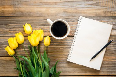 Boş bir defter, bir fincan sade kahve ve ahşap arka planda sarı laleler..