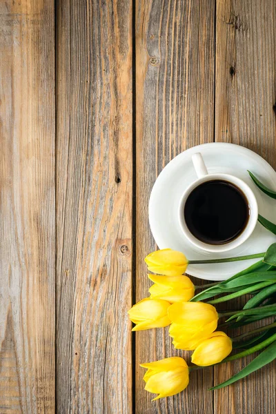 stock image Bouquet of bright yellow tulips and cup of black coffee on a wooden background, top view, copy space.