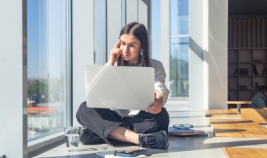 Modern bir ofiste, bir yaz sabahı, pencerenin yanındaki dizüstü bilgisayarın önünde düşünceli genç bir kadın..
