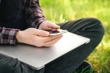 Adamın biri elinde dizüstü bilgisayar ve akıllı telefonla çimlerin üzerinde oturuyor. Açık alanda, yakın plan cihazlar kullanıyor..