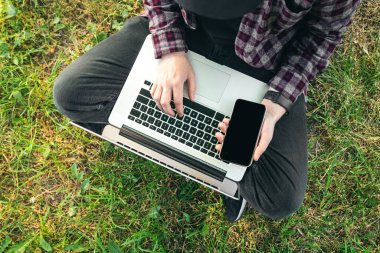 Parkta çimlerin üzerinde oturan bir adam dizüstü bilgisayarın başında oturur ve çalışır. Akıllı telefonu eliyle tutar..