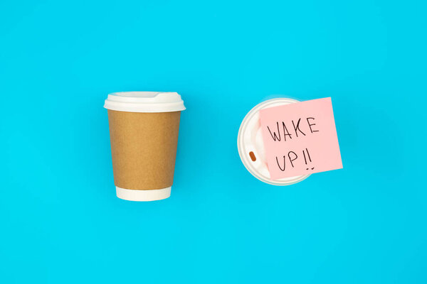 Paper disposable cups and paper reminder with text Wake up on blue background, top view.