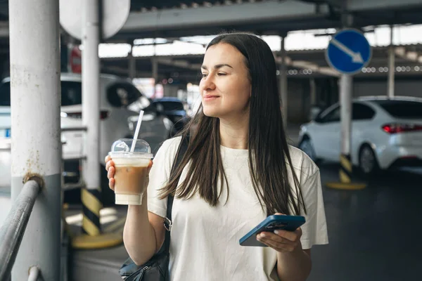 Young woman with coffee and smartphone in the parking lot, woman waiting for a taxi in the parking lot.