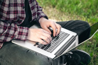 Parkta elinde dizüstü bilgisayarla çimlerin üzerinde oturan bir adam. Kapalı elleri ve laptopu var. Yazın dışarıda çalışıyor..