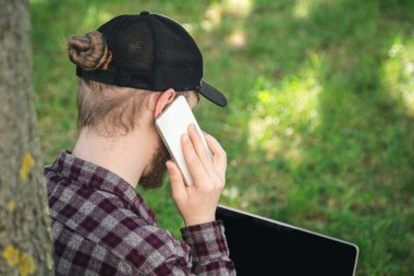 Yakın plan, akıllı telefondan konuşan bir adam çimlerin üzerinde dizüstü bilgisayarla oturuyor, dışarıda çalışıyor..