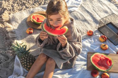 A little girl on a sandy seashore on a blanket at a picnic with fruits eats watermelon.