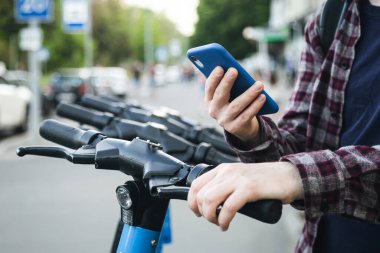Elinde cep telefonu olan bir adam açık hava gezisine hazırlanırken elektrikli scooterın kilidini açıyor..