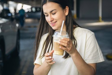 Otoparkta tek kullanımlık bardakta kahve içen genç bir kadın, arabayla yolculuk, turizm ve şehir yaşam tarzı konsepti.