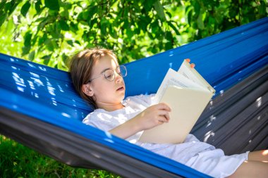 Gözlüklü tatlı genç kız bahçede bir hamakta yatarken kitap okuyor..