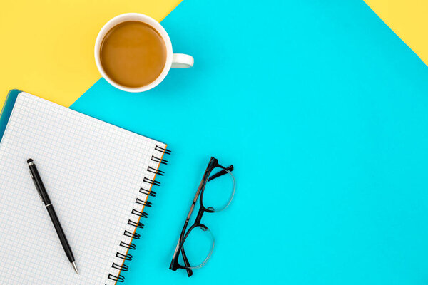 Flat lay design of work desk with notebook, eye glasses and cup of coffee on a blue and yellow background.