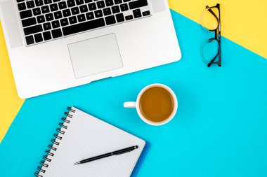 Not defteri, dizüstü bilgisayar, gözlük ve kahve fincanı mavi ve sarı arka planda, düz bir çalışma masası dizaynı.