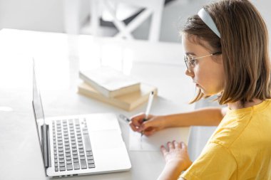 Ev ödevi yapan genç bir kız kitap ve dizüstü bilgisayarla oturuyor, online öğrenim kavramı, evde okuyan bir çocuk..