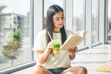 Çekici genç bir kadın kitap okuyor ve pencerenin yanında otururken soğuk bir içecek içiyor..