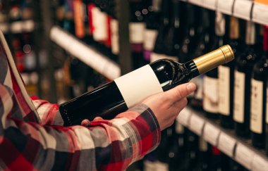 Şarap dükkanında elinde şarap şişesi tutan bir adam raflardaki şarap şişelerinin çeşitlerinden doğru şarabı seçiyor..