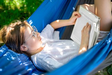 Gözlüklü tatlı genç kız bahçede bir hamakta yatarken kitap okuyor..