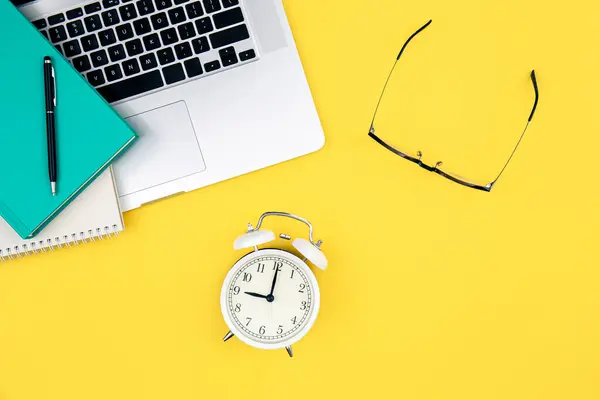 Laptop, alarm clock and glasses on a yellow background, top view.