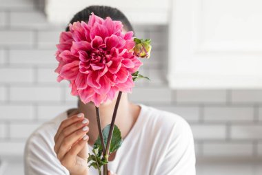 Bir kadın beyaz mutfak arka planında büyük pembe bir Dahlia çiçeğiyle yüzünü kapatıyor..