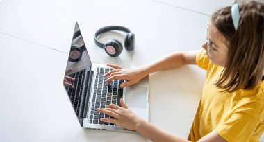 Gözlüklü bir genç kız evde bir masada otururken dizüstü bilgisayar kullanıyor, bir çocuk da çalışmak ve eğlenmek için bilgisayar kullanıyor..
