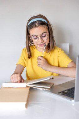 Gözlüklü ve sarı tişörtlü genç bir kız, kitaplarla ve laptopla ödev yapıyor, odada bir masada oturuyor..