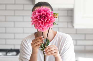 Bir kadın beyaz mutfak arka planında büyük pembe bir Dahlia çiçeğiyle yüzünü kapatıyor..