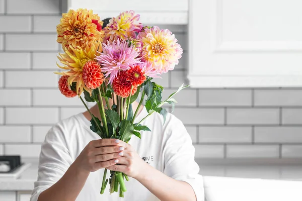 A woman holding a bouquet of bouquet of dahlia flowers a light kitchen interior background, copy space.