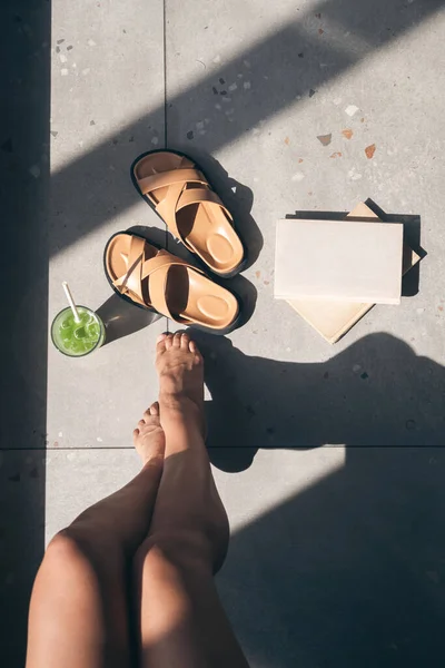 Summer composition with slippers, lemonade, book and womens feet on a tile with shadows on a sunny day, top view.
