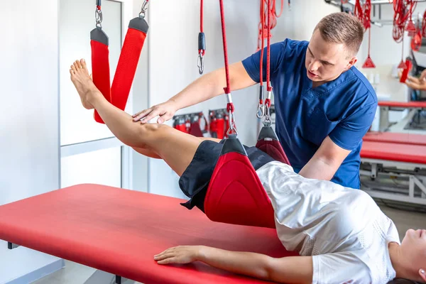 stock image A young female is doing exercises with special ropes in rehabilitation center, physiotherapist supports his client during workout to recover after the injury.