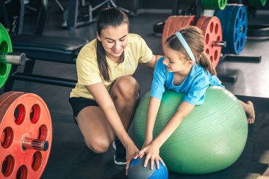 Küçük sporcu kız ve genç güzel anne spor salonunda spor topuyla esneme hareketleri yapıyorlar..