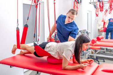 Genç bir kadın rehabilitasyon merkezinde özel iplerle egzersiz yapıyor, fizyoterapist sakatlıktan sonra iyileşmek için antrenman sırasında müvekkiline destek oluyor..