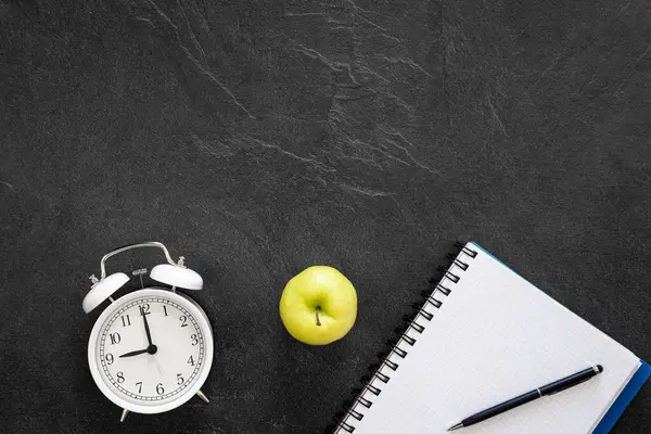 Alarm clock, notepad and apple on black background, top view, school concept, copy space.
