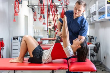 Kadın hastaya süspansiyon kayışları tedavisi uygulayan rehabilitasyon uzmanı, ağrısız dolaşım düzenini iyileştirmek için terapi egzersizleri, nöro-fonksiyon teknikleri..