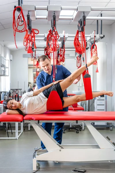 Female patient hanging on suspensions at rehabilitation center, therapeutic exercises and neuromuscular activation on red cord slings, Neurac technique.
