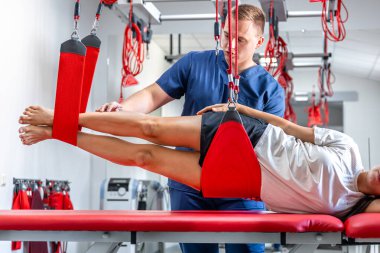 Genç bir kadın rehabilitasyon merkezinde özel iplerle egzersiz yapıyor, fizyoterapist sakatlıktan sonra iyileşmek için antrenman sırasında müvekkiline destek oluyor..