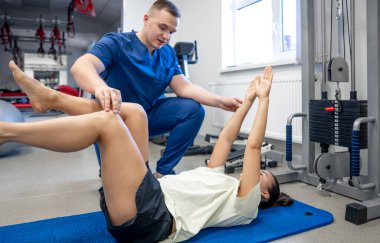 Woman laying at the yoga mat and doing gymnastics with help of his young physical therapist. clipart