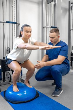 Fizyoterapist kadın hastaya rehabilitasyonda yardım ediyor, bosu topuyla denge egzersizi yapıyor, kadın ve doktor spor salonunda fizik tedavi görüyor..