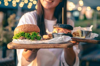 Elinde iki hamburger tutan çekici genç bir kadın, arka planda bulanık bir şekilde sokak kafesinde fast food yiyor..