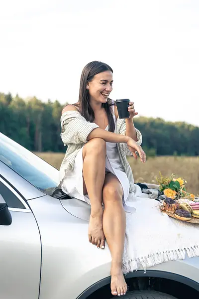 Attractive young woman sits on a car and drinks coffee in a field, enjoy a summer weekend picnic with a car outside the city in a field on a sunny day, sunset, vacation and trip.